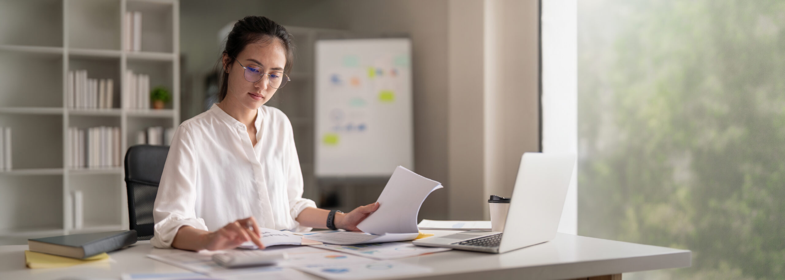 "A focused Asian woman wearing glasses and a white shirt, working at a desk with documents, a calculator, and a laptop in an office setting