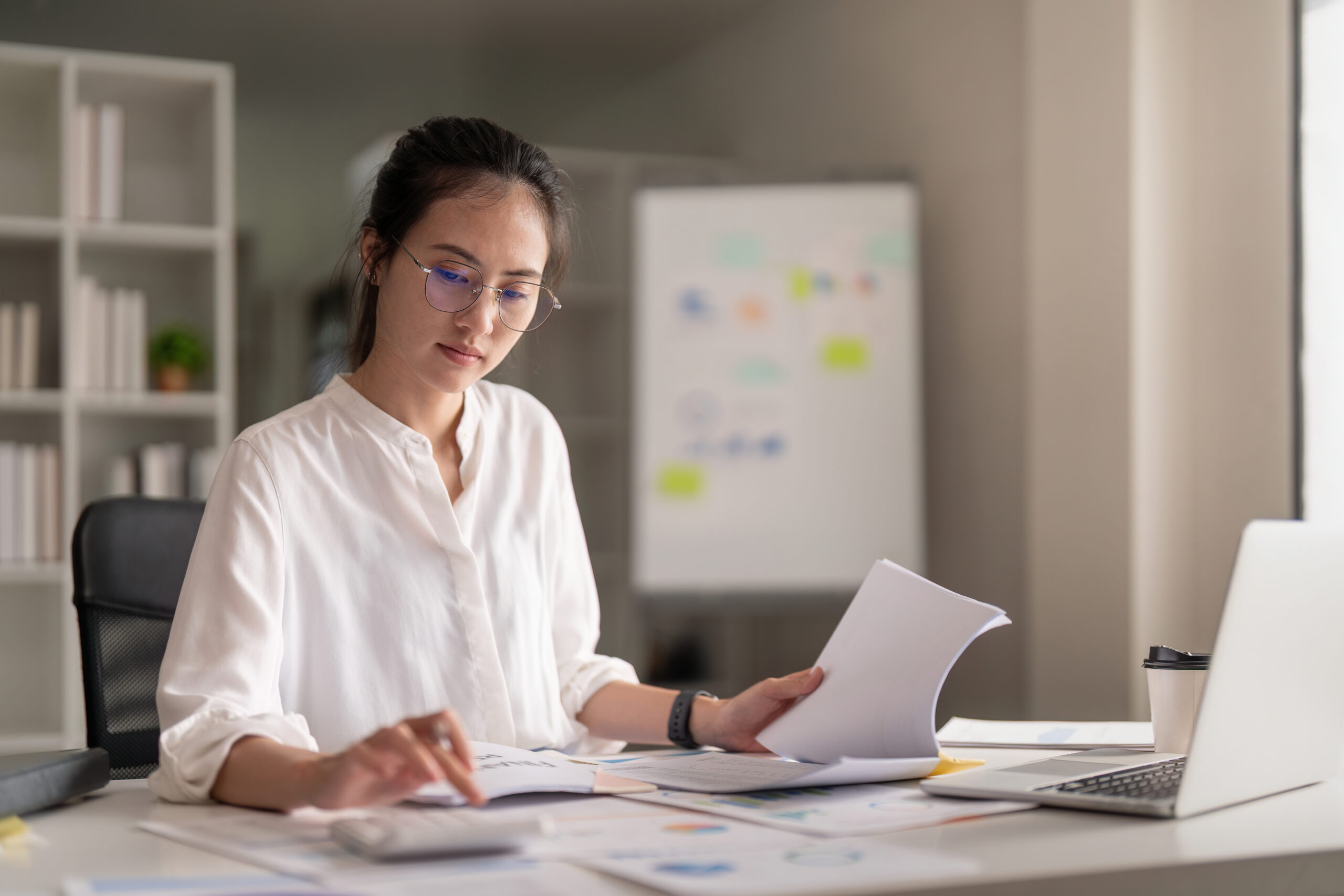 "A focused Asian woman wearing glasses and a white shirt, working at a desk with documents, a calculator, and a laptop in an office setting