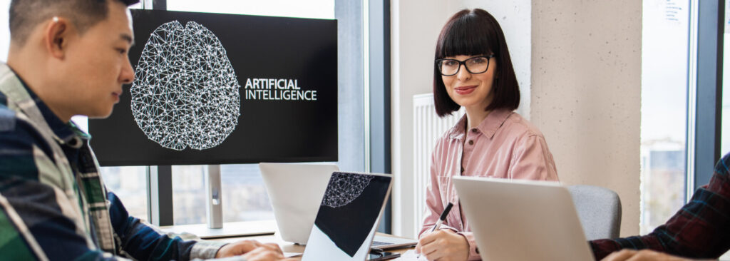 Eine kollaborative Büroszene mit drei Personen, die an Laptops arbeiten, zentriert um einen großen Monitor, der eine polygonale Gehirngrafik und die Worte „ARTIFICIAL INTELLIGENCE“ zeigt. Die Umgebung spiegelt Innovation und Teamarbeit in der Technologie- und KI-Branche wider.