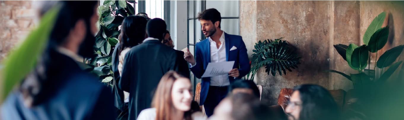 A man in a blue suit speaks to colleagues during a meeting in a modern office with large plants and industrial-style windows.