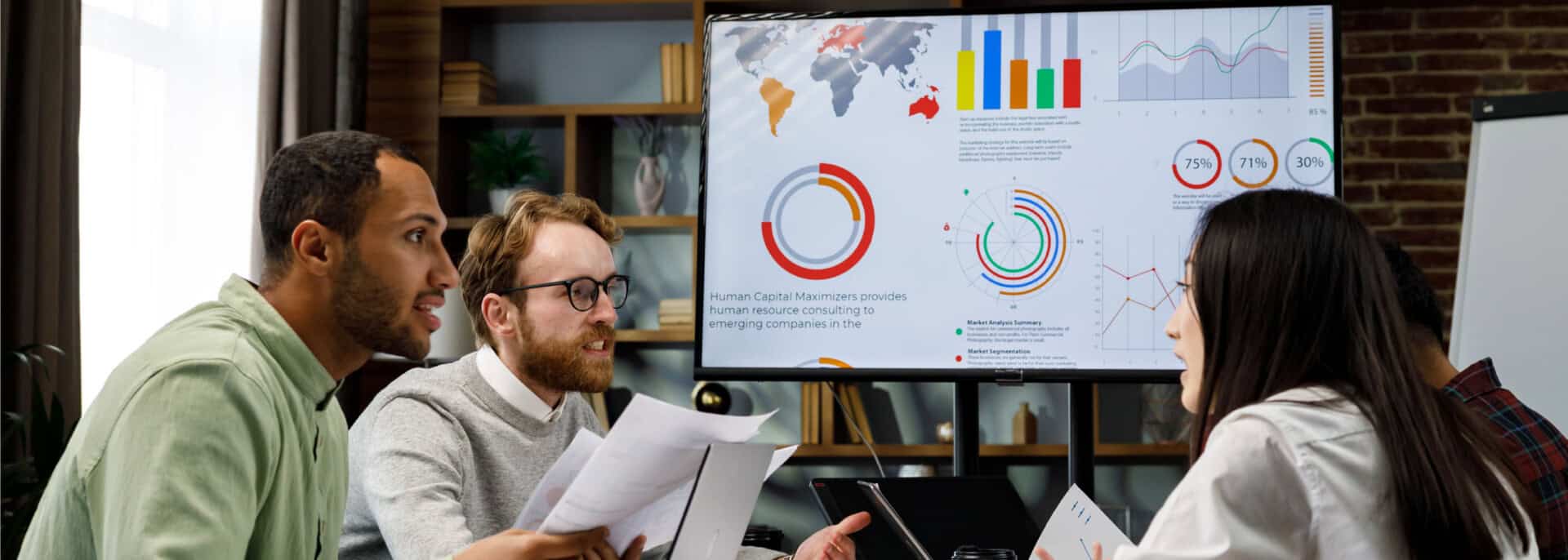 Three professionals reviewing charts, analytics, and printed reports on a large screen during a business strategy meeting in a modern office.
