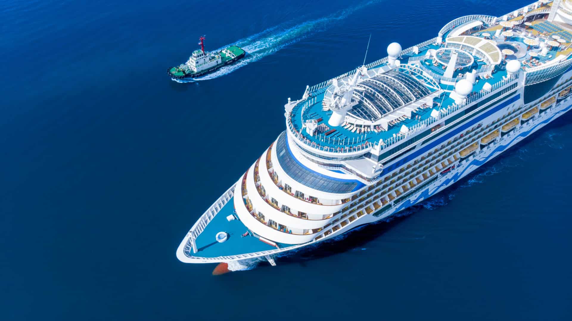 Aerial view of a large white luxury cruise ship with blue accents navigating deep blue waters, accompanied by a smaller tugboat trailing behind. The cruise ship features tiered decks, a pool area, lounge chairs, and vibrant yellow umbrellas, with tiny figures visible on the decks. The scene is captured from a high-angle perspective, emphasizing the ship's scale and the vastness of the ocean.
