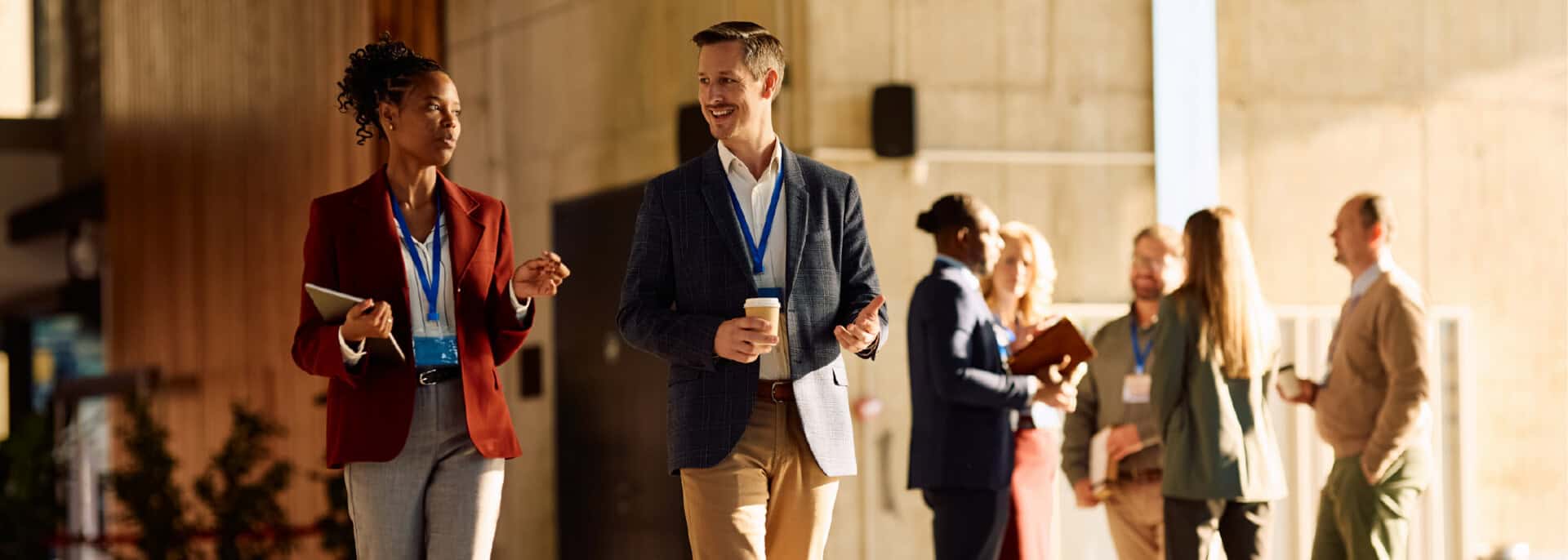 Two business professionals walk and talk in a sunlit office lobby while other colleagues network in the background.