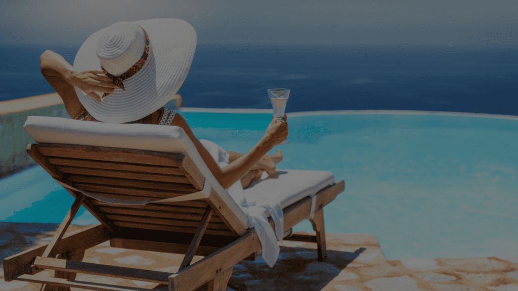 Woman relaxing on a poolside lounge chair, holding a glass of champagne and looking over an infinity pool toward the ocean.