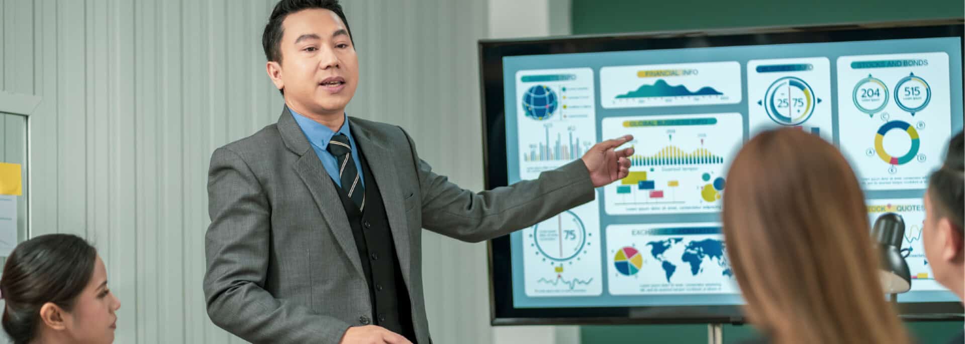 Business presenter in a grey suit points to a hotel business intelligence dashboard with financial charts, global maps, and performance metrics as an audience looks on.