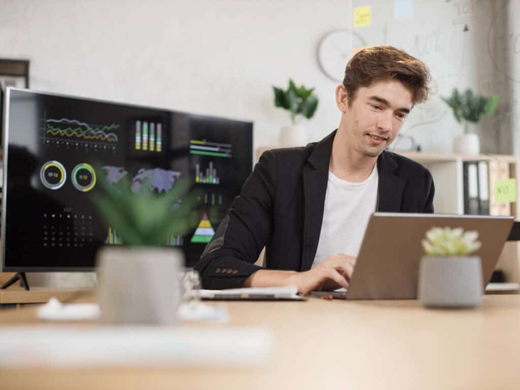 Professional working on a laptop in an office, with charts and a world map displayed on a screen behind him.